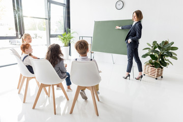 young female teacher holding chalk and pointing at blackboard while explaining lesson to students