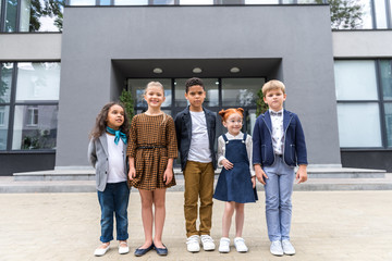 group of adorable multiethnic schoolchildren smiling at camera while standing near school