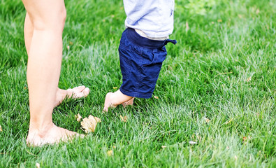 Little child doing First steps on green grass. Legs of young mother and her baby son