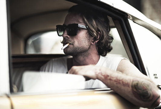 Man with moustache sitting in car, wearing sunglasses, smoking a cigarette, tattooed arm leaning out of window.