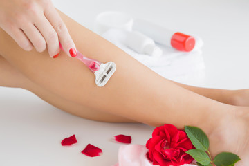 Fototapeta premium Young woman removing hair on legs with razor. Shaving every day. Smooth skin. Body care with soft cream. Summer atmosphere with fresh and fragrant red rose.