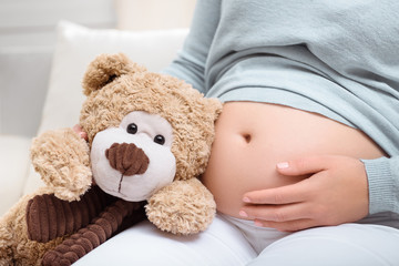 cropped view of pregnant woman with teddy bear near her belly at home