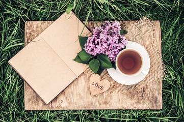 A cup of black tea and an open diary with blank pages on green grass. Romantic concept. Wooden card with a heart. Vintage tinting