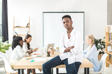 portrait of pensive african american businessman with arms crossed sitting on table with multicultural colleagues working behind