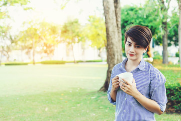 Beautiful short hair woman standing in the green yard and drinking a favorite beverage in white cup for resting between her outdoor working business in the park.