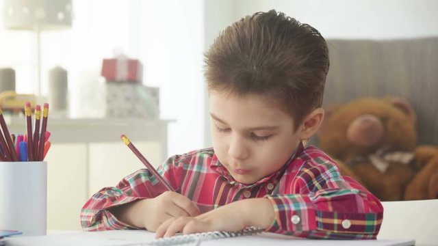 small kid boy draw with crayons and color pictures on paper with enthusiasm sitting at the table at home