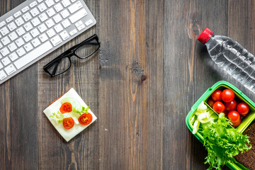 Eat at workplace. Lunch box with vegetable sandwiches near computer on dark wooden background top view copyspace
