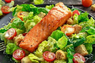Caesar Salmon Salad with croutons, parmesan cheese, tomatoes, dressing and pepper in a black plate on wooden table.