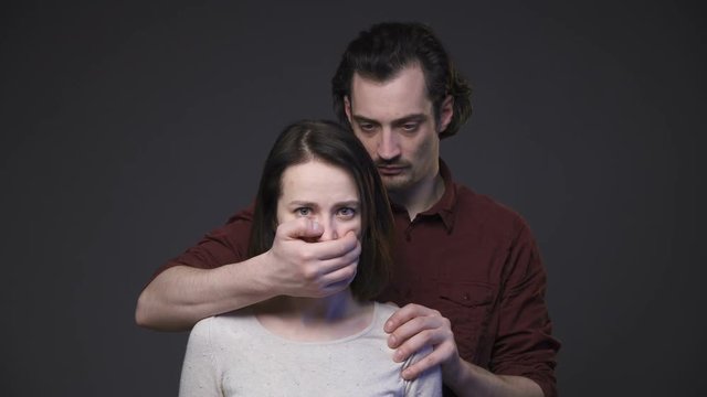 Home violence, man closing woman's mouth by the hand, gray background