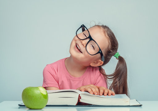 cheerful beautiful cute little girl next to thick book and green apple looking at camera