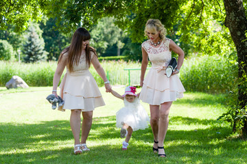 Obraz premium Funny baby girl in white dress is playing with mother and godmother in the garden at christening