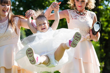 Funny baby girl in white dress is playing with mother and godmother in the garden at christening