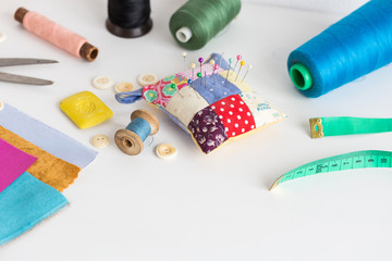 sewing tools, patchwork, tailoring and fashion concept - close-up on white desk in studio, pincushion, thread spools, measuring meter, buttons, scissors, soap, pieces of colored patchwork fabric.
