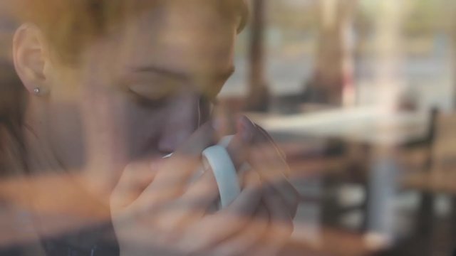 Beautiful young girl sits in a cafe near a window and drinks hot black coffee. The glare of the street is displayed on the glass.