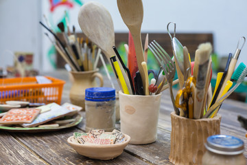 Tools and accessories for manual work. Blurred paint brushes in the jar. Clay cups on a wooden table. Accessories and tools in the creative workshop.