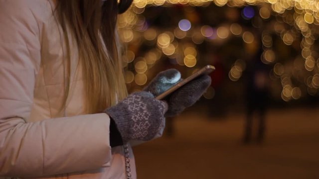 The girl uses the touch screen on smartphone in winter against the background of the night lights of the Christmas, New Year, multi-colored Christmas tree.