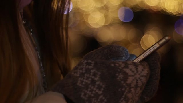 The girl uses the touch screen on smartphone in winter against the background of the night lights of the Christmas, New Year, multi-colored Christmas tree.