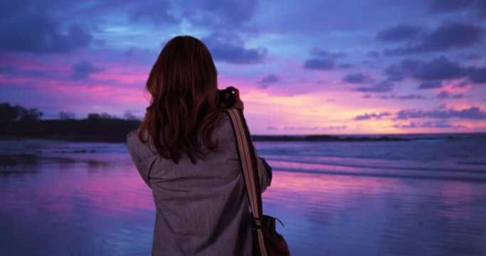 Female traveler standing on beach taking picture of calm ocean at sunset