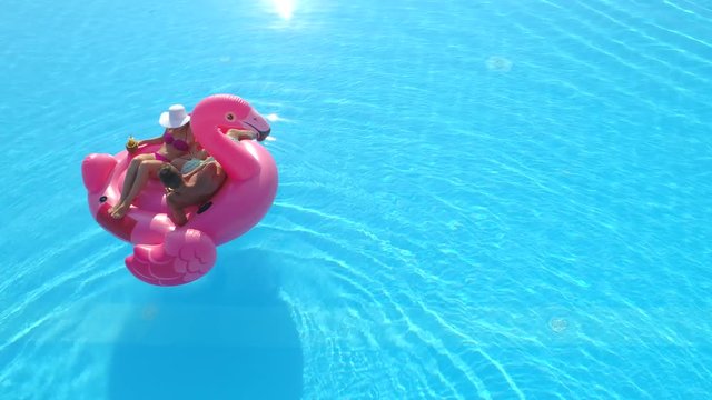 AERIAL: Young couple drinking cocktails and relaxing while lying on fun inflatable flamingo floatie. Sexy couple in swimsuit sipping drinks and enjoying on pink flamingo floating on sunny pool water