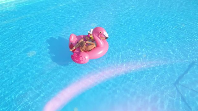 AERIAL LENS FLARE: Relaxed couple lying on fun inflatable flamingo floatie, drinking cocktails and relaxing. Sexy couple in swimsuit sipping drinks and enjoying on pink flamingo floating in sunny pool