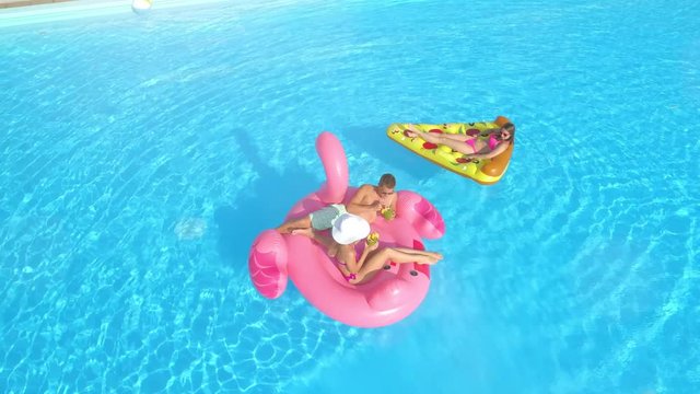 AERIAL LENS FLARE: Young fit girls and guy in swimsuits laying on fun inflatable pizza and pink flamingo floating on water. Happy friends on vacation relaxing on pink flamingo and pizza floats in pool