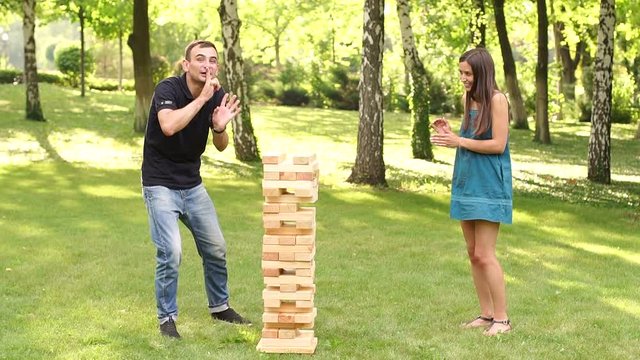 Two cheerful young people playing big jenga wooden blocks in the Park on the grass, the jenga tower falls. Slow motion.
