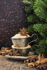 Obraz na płótnie Canvas Melchior vintage cups with cane sugar, anise stars, Indian Indian cinnamon sticks on an aged wooden background surrounded by spruce spruce branches. with imitation of falling snow
