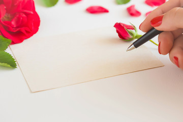 Woman's hand with pen writing on the white blank greeting card on the table. Red roses. Empty place for a text.