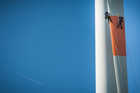 Inspection engineers abseiling down a rotor blade of a wind turbine in a North German wind farm on a clear day with blue sky