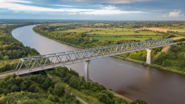 Bridge crossing the Kiel Canal (German: Nord-Ostsee-Kanal) in Schleswig Holstein in Germany with lush green fields and wind farms in the background is captured aerial from a drone at sunset.