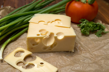 mouthwatering delicious piece of maasdam cheese and a slice on a wooden table surrounded by greenery and tomatoes