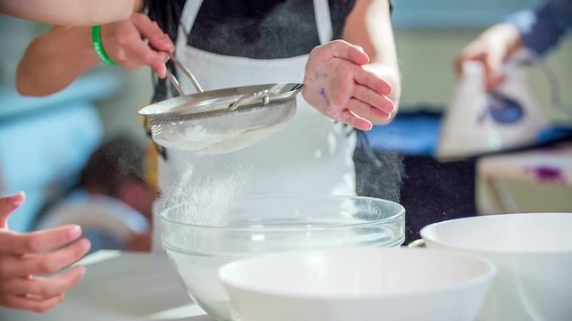 Students in class are putting a lot of flour on a sifter and then they are sprinkling it through a sifter.