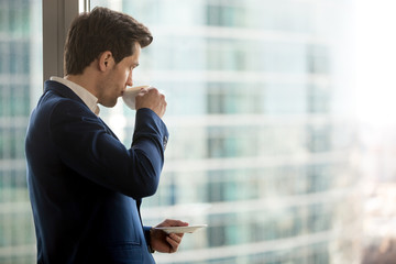 Thoughtful businessman drinking coffee, looking through window at big modern city, having break, deep in thoughts, enjoying view, waiting for meeting to start, making business decision, copy space
