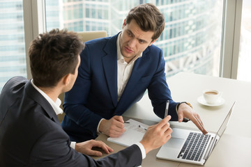 Two serious businessmen using laptop and discussing new project at office, developing strategy for online business, explaining sharing ideas, preparing presentation, having brainstorming session