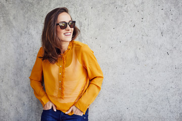 Cheerful young woman leaning against concrete wall with hands in pockets