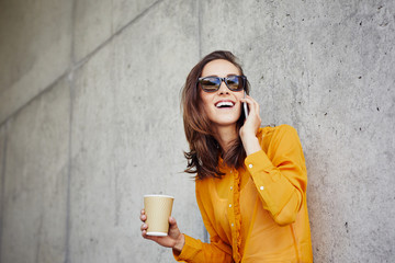 Cheerful young woman talking on phone and holding coffee while leaning against wall outside