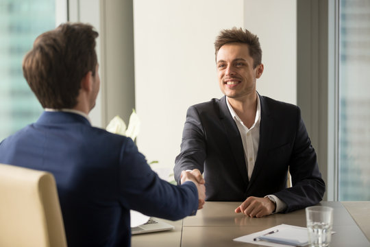 Two cheerful businessmen shaking hands over office table, starting negotiations, hr greeting applicant arrived at job interview with handshake, good first impression, forming beneficial partnership
