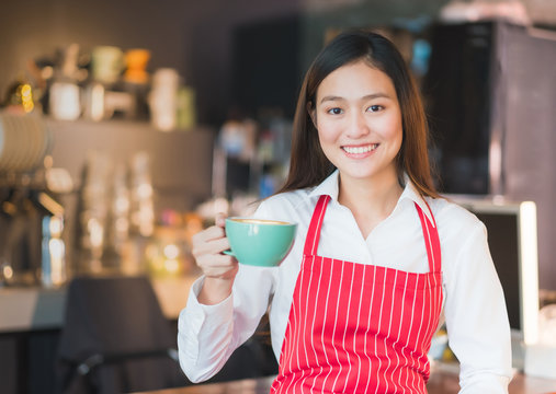 Asian female barista wear red apron holding hot cappuccino coffee cup menu at counter bar with smile face,cafe service concept