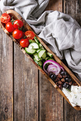 Ingredients for traditional greek salad. Cherry tomatoes, sliced cucumbers, red onion, black olives, feta cheese in olive wood bowl with gray textile over wooden plank background. Top view
