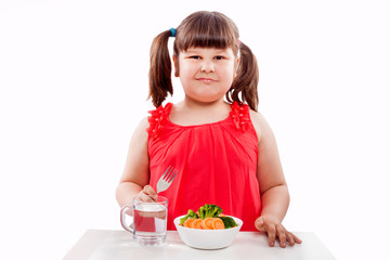 Girl likes healthy food, sits at the table near the plate with vegetables. Child isolated on white background