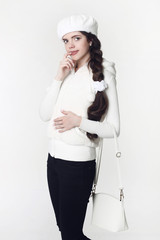 Fashion teen white style look. Fashionable young girl wears fur jacket and beret, holding handbag, posing isolated on studio background.