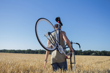 young hipster in sunglasses walk in the fields with bicycle on shoulder, fields and blue sky background 
