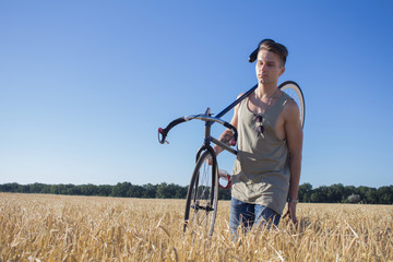 Naklejka na ściany i meble young hipster in sunglasses walk in the fields with bicycle on shoulder, fields and blue sky background 