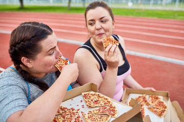 Happy young over-size women eating tasty and appetizing pizza after workout on stadium