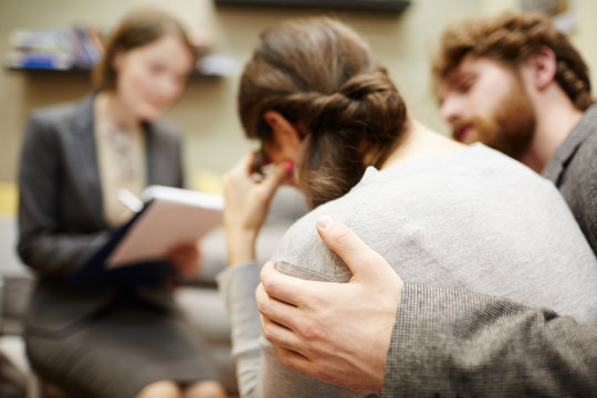 Back view image of young woman crying in couples counseling session with  husband hugging her supportively