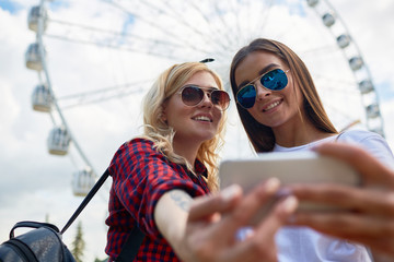 Portrait of two beautiful young girls taking selfie via smartphone camera standing against tall...