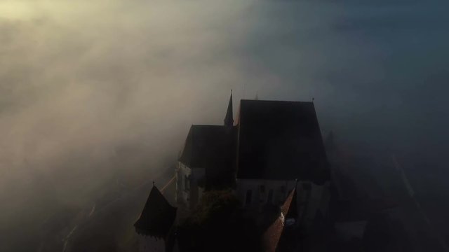 4k aerial orbit flight around medieval church in saxon village Biertan.	Impressive lutheran church appearing through clouds in early morning. Transylvania, Romania.