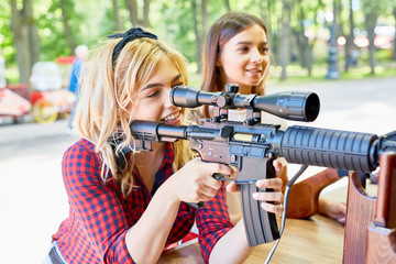 Portrait of beautiful blond woman aiming with big rifle at outdoor game tent in amusement park