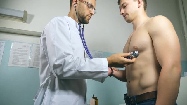 Doctor examining young male patient with stethoscope. Medic checking chest of guy in her office at the hospital. Medical worker listening heartbeat of athletic man. Close up