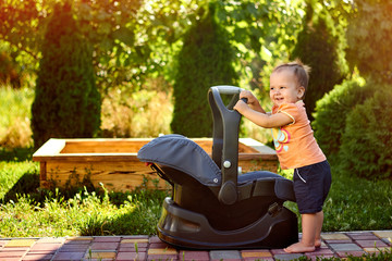Infant baby standing near modern car seat in a park. New born child traveling by car. Child safety on the road. Safe way to travel with young kids. Fastened seat belts. Trip with an infant.
